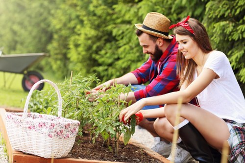 A young man and woman are gardening outdoors in a landscaped garden in Palmers Green, with lush green trees and shrubs in the background. They are both kneeling beside a freshly prepared flower bed or vegetable patch, tending to the plants. The man, wearing a straw hat and a red plaid shirt, is examining or pruning the greenery, while the woman, dressed in a white t-shirt with a red headband, is also reaching into the soil, planting or weeding. To their left, a large white wicker basket with a floral pattern rests on the edge of the garden bed, likely used for collecting garden waste or harvest. The garden features neat, well-maintained soil, with a variety of healthy, leafy plants growing in the bed. The overall environment appears bright and mild, suggesting a sunny day suitable for outdoor gardening activities, which reflect professional gardening practices supported by Landscapers Palmers Green working within local outdoor spaces and focused on sustainable gardening and recycling initiatives.