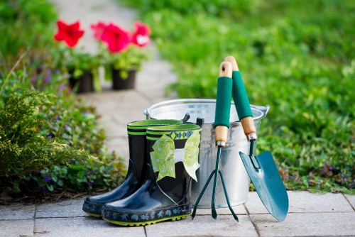 This image depicts a section of a well-maintained garden pathway edged with lush green grass, bordered by flowering plants and shrubs. In the foreground, there is a pair of black rubber gardening boots with green leaf-patterned gaiters draped over the top, suggesting outdoor footwear used for gardening tasks. Next to the boots, a white plastic bucket is visible, partially filled with gardening tools, including a small hand trowel with a teal handle and a hand fork. Adjacent to the bucket, a metal garden spade with a teal handle is resting on the ground. The background features a paved stone pathway, leading through the garden, with potted flowers in bloom, including vibrant red and pink blossoms, indicating a cultivated, colorful outdoor space typical of a residential garden in Palmers Green, London. The scene suggests active gardening or recent outdoor maintenance, with natural daylight illuminating the details. This setting ties into gardening services provided by Landscapers Palmers Green, emphasizing outdoor landscaping, lawn care, and garden maintenance in the local area.