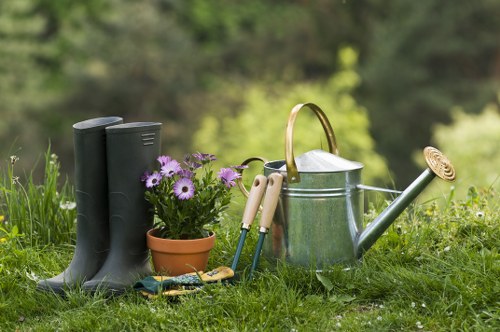 In a lush, green outdoor garden space, there are a pair of black rubber gardening boots standing upright on the grass, with a small terracotta pot containing purple and white flowers placed nearby. A metal watering can with a wooden handle and a long spout is positioned on the grass, accompanied by gardening tools, including a pair of hand trowels and pruning shears, laid out in front of the tools. The background features blurred trees and foliage, indicating a natural outdoor environment suitable for gardening and landscape maintenance. The scene is well-lit with natural daylight, capturing a peaceful moment of garden preparation or plant care in a typical outdoor garden area, aligning with gardening services offered by Landscapers Palmers Green in the local area, possibly around the N13 postcode, enhancing the visual connection to local landscaping and lawn care work.
