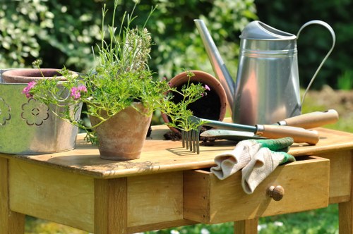 A wooden outdoor garden table situated in a lush, green garden with various gardening tools and plant pots arranged on top. The table displays a terracotta pot with blooming pink flowers and a larger pot with green, leafy herbs or plants. A metal watering can with a curved handle is positioned to the right, alongside a pair of gardening gloves and small hand tools such as a trowel and a hand rake. The surrounding environment features vibrant green foliage and a well-maintained lawn, indicating a neatly cared-for outdoor space suited for gardening activities. This scene reflects typical garden maintenance with a focus on plant care and outdoor landscaping, relevant to services offered by Landscapers Palmers Green in the local garden and lawn care sector, within the context of a well-cared-for residential garden in the UK.