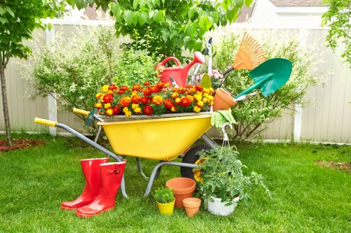 A brightly colored garden scene featuring a yellow wheelbarrow filled with vibrant red and yellow marigold flowers, situated on a well-maintained green lawn. The wheelbarrow has a red watering can inside and a turquoise garden fork and orange watering can resting against it. Nearby, a pair of red rubber wellington boots are placed on the grass, alongside small terracotta plant pots and a white pot with blooming green foliage. In the background, a wooden fence encloses the garden, with lush green trees and shrubs providing a shaded, natural backdrop. The overall environment appears well-cared for, with the garden scene suggesting ongoing outdoor maintenance and planting activity typical of gardening services in Palmers Green or the surrounding areas in North London, supported by professional landscapers and garden care providers.