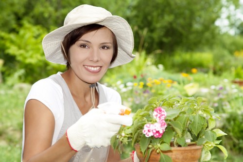 The image depicts a woman in a garden with lush green foliage and flowering plants, including a potted plant with pink blossoms that she is tending to. She is wearing a wide-brimmed white hat, a white short-sleeved top, and gardening gloves, smiling as she carefully examines or plants the flowers. The garden background features a variety of plants, grass, and possibly flowerbeds, suggesting a well-maintained outdoor space suited for gardening and landscaping activities. The natural lighting indicates a bright day, contributing to the vibrant green and floral colours. This scene exemplifies outdoor gardening work typical of professional gardening services in Palmers Green, and it illustrates the importance of attentive planting and garden maintenance, aligning with the landscaping skills offered by Landscapers Palmers Green on their Planting Services page.