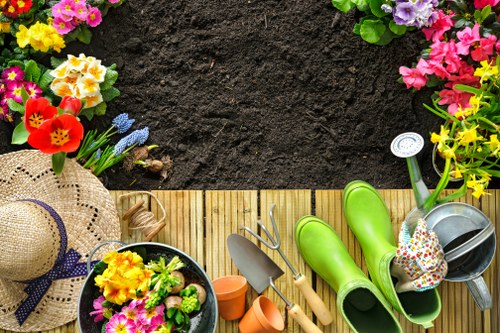 A top-down view of a garden area featuring a section of dark, rich soil bordered by a wooden decking platform. The garden is decorated with vibrant flower beds containing yellow, pink, purple, and red flowering plants and shrubs, creating colorful borders around the planting area. On the wooden surface, there are gardening tools including a small trowel, a pair of scissors, and a watering can, along with bright green rubber boots, a straw sun hat with a blue ribbon, and a small black pot filled with pink and yellow flowers. The background shows lush green foliage and additional flower arrangements, indicating a well-maintained outdoor space suitable for gardening and landscaping services. The scene is evenly lit, suggesting a clear, sunny day, emphasizing the natural tones and textures of the garden environment and outdoor maintenance equipment.