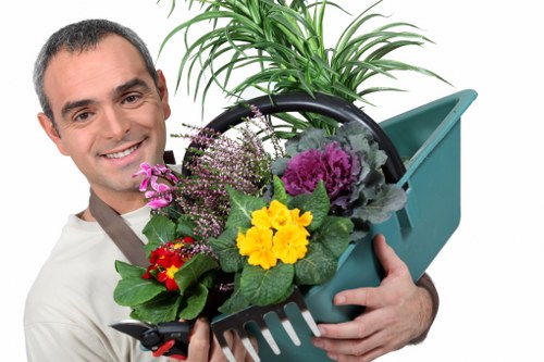 A smiling man with short dark hair and a light beige shirt holds a teal garden watering can filled with various colourful flowering plants and green foliage. The plants include purple asters, yellow primroses, red flowers, and leafy greens, arranged in a decorative manner. Behind him, a large potted ornamental grass with long, slender green blades is visible, positioned on a paved surface in a garden environment. The background suggests an outdoor space with soft natural lighting, indicative of a bright, slightly cloudy day. The setting appears to be a landscaped garden, highlighting elements such as flower beds, paving, and lush greenery, which are typical features of a well-maintained outdoor area in Palmers Green and relevant to local gardening services provided by Landscapers Palmers Green.