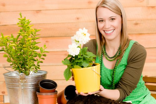 The image shows a woman with blonde hair smiling as she holds a bright yellow flowerpot containing white flowering plants, likely geraniums or similar flowering shrubs, in a garden or outdoor workspace. She is seated at a wooden table or work surface, surrounded by gardening tools and materials including a metal bucket with a green shrub and several empty plant pots of various sizes and colours, such as black, terracotta, and dark brown. In the background, there is a wooden fence or paneling, providing a natural and warm setting typical of a well-maintained garden or landscaped yard. The lighting suggests a bright, cloudy day, enhancing the natural tones of the garden elements. This scene exemplifies gardening activities like planting or potting, aligning with services offered by Landscapers Palmers Green, specialising in gardening and outdoor maintenance within the local area, possibly near N13, Palmers Green, or surroundings. The arrangement of healthy plants and gardening tools reflects ongoing garden care aimed at enhancing outdoor environments in residential gardens or landscaped front yards.