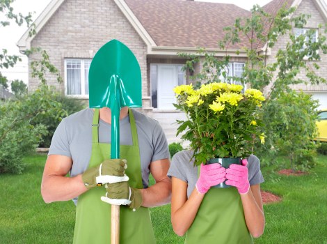 A person wearing a green gardening apron and yellow gloves is holding a small shovel, standing beside a person with a pink gardening glove, who is holding a vibrant yellow flowering plant in a pink container. The scene is set in a residential garden with a well-maintained green lawn, and young trees and shrubs surround a light-colored brick house with white-framed windows and a tiled roof in the background. The garden features smooth grass, neatly arranged flower beds, and a paved pathway visible near the house. The weather appears bright and sunny, casting natural light across the scene, which highlights the healthy foliage, the blooming flowers, and the outdoor environment typical of gardening services in Palmers Green or nearby areas. This image is relevant to gardening and landscaping services offered by Landscapers Palmers Green, emphasizing garden planting and maintenance activities in a suburban backyard setting.