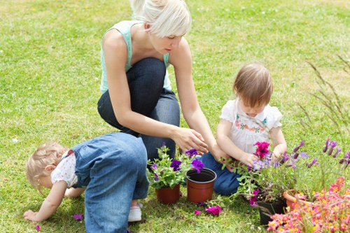 A woman with short blonde hair, wearing a light blue sleeveless top and dark jeans, is kneeling on a well-maintained grassy lawn in a front garden, tending to a flower bed alongside two young children. One child, dressed in blue overalls, is bent over, examining or picking flowers or foliage. The other child, wearing a white top with floral patterns, is sitting near the woman, holding a small cluster of pink flowers. The flower bed features a variety of colorful blooms, including purple and pink flowers, with some in black plant pots and others directly in the soil, bordered by a neat edge separating it from the lush green lawn. The background shows a tidy garden setting with a hedge or shrub line, creating a natural boundary. The scene suggests outdoor garden maintenance or planting activity on a bright, possibly sunny day, with natural daylight illuminating the scene. This image captures a moment of family gardening, typical of local outdoor space improvements and flower bed ideas in Palmers Green, highlighting garden elements such as soil, flowers, and lawn care suitable for gardening services in the area.