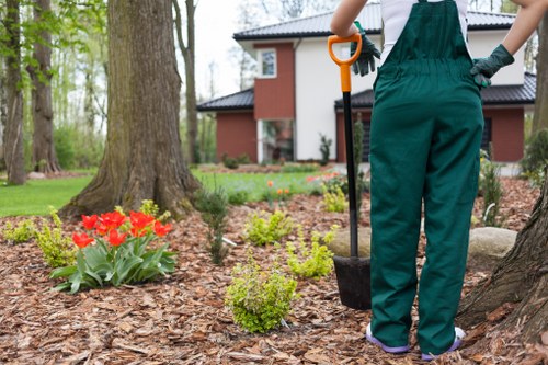 A gardener in green overalls using a shovel to prepare a flower bed in a well-maintained front garden in Palmers Green, North London. The garden features a mature tree with a textured trunk, surrounded by a variety of plants including red tulips and low-growing green shrubs planted in mulched soil. In the background, a modern two-storey house with a red brick exterior, white window frames, and a black tiled roof is visible, with a lush green lawn extending around the area. The scene is set on a bright, slightly overcast day, with natural lighting highlighting the vibrant colors of the flowers and the healthy foliage. This outdoor space reflects careful landscaping where professional gardening services help maintain the neat flower beds, lawn, and trees, supporting a tidy and inviting garden environment.
