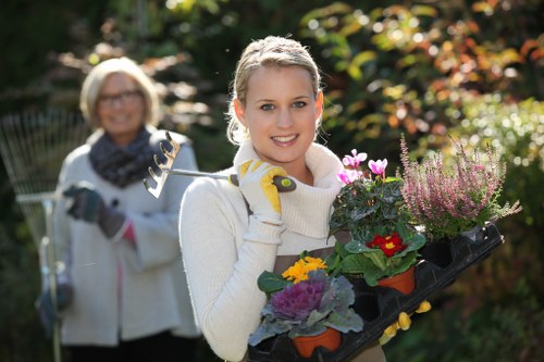 A young woman with blonde hair, wearing a cream sweater and gardening gloves, is smiling and holding a black tray filled with colorful flowering plants, including petunias and heathers, in a well-maintained garden during daylight. Behind her, an older woman with glasses and a light jacket is slightly out of focus, also smiling and holding garden tools, with lush green trees and shrubs in the background, suggesting a healthy, landscaped outdoor space typical of residential gardens in Palmers Green. The garden features a mix of vibrant plant colors, natural soil patches, and paved pathways, indicating regular outdoor maintenance and gardening care. The weather appears sunny and clear, ideal for gardening activities, reflecting professional gardening services by Landscapers Palmers Green which focus on outdoor garden design, planting, and maintenance.