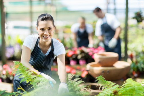 A smiling woman wearing a striped apron and gardening gloves is shown working in a lush greenhouse or garden centre, surrounded by a variety of potted plants and flowering shrubs. She is leaning slightly forward, tending to green fern-like foliage in the foreground, with her hands engaged in planting or adjusting the plants. Behind her, other staff members in similar uniforms are visible, attending to additional plants or arrangements, indicating a team effort in horticultural care. The environment features a bright, naturally lit space with glass panels allowing sunlight to illuminate the greenery, showcasing a vibrant and well-maintained outdoor or botanical setting. The scene emphasizes healthy, vivid green and pink tones in the foliage and flowers, with textured soil, wooden or tiled surfaces, and organized plant displays. This outdoor or indoor gardening activity aligns with professional landscaping and gardening services, such as those offered by Landscapers Palmers Green, catering to garden maintenance, planting, and landscape enhancement in the local area around N13 or Palmers Green.